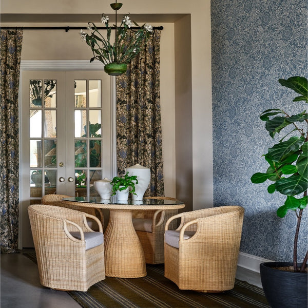 A dining area featuring a round glass-top table with a woven natural rattan pedestal base, paired with two matching woven armchairs with light cushions. The room has a textured blue-gray wall, patterned floral drapes, and a potted green plant.
