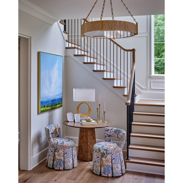 A styled interior entryway with a wooden staircase, a round wood table, two patterned upholstered chairs, a table lamp, and a large framed landscape painting on the wall.