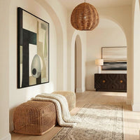 A styled interior hallway with cream walls, arched alcoves, a woven rattan pendant light, a matching woven bench with a cream knit throw, a patterned area rug, and framed abstract wall art.