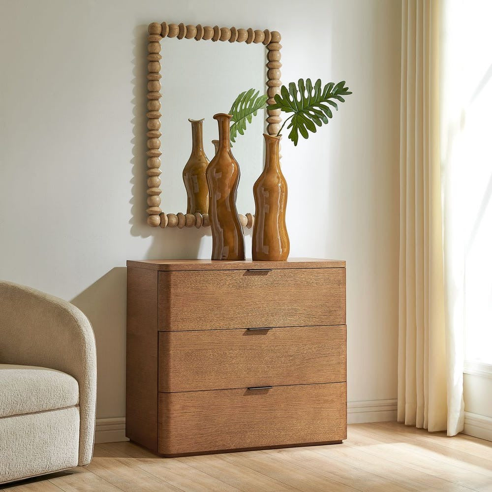 A three-drawer wooden chest of drawers in a warm medium brown finish, styled in a bright room with a matching wood beaded mirror, decorative vases, and a curtain.