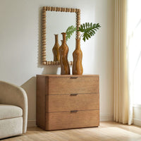 A three-drawer wooden chest of drawers in a warm medium brown finish, styled in a bright room with a matching wood beaded mirror, decorative vases, and a curtain.