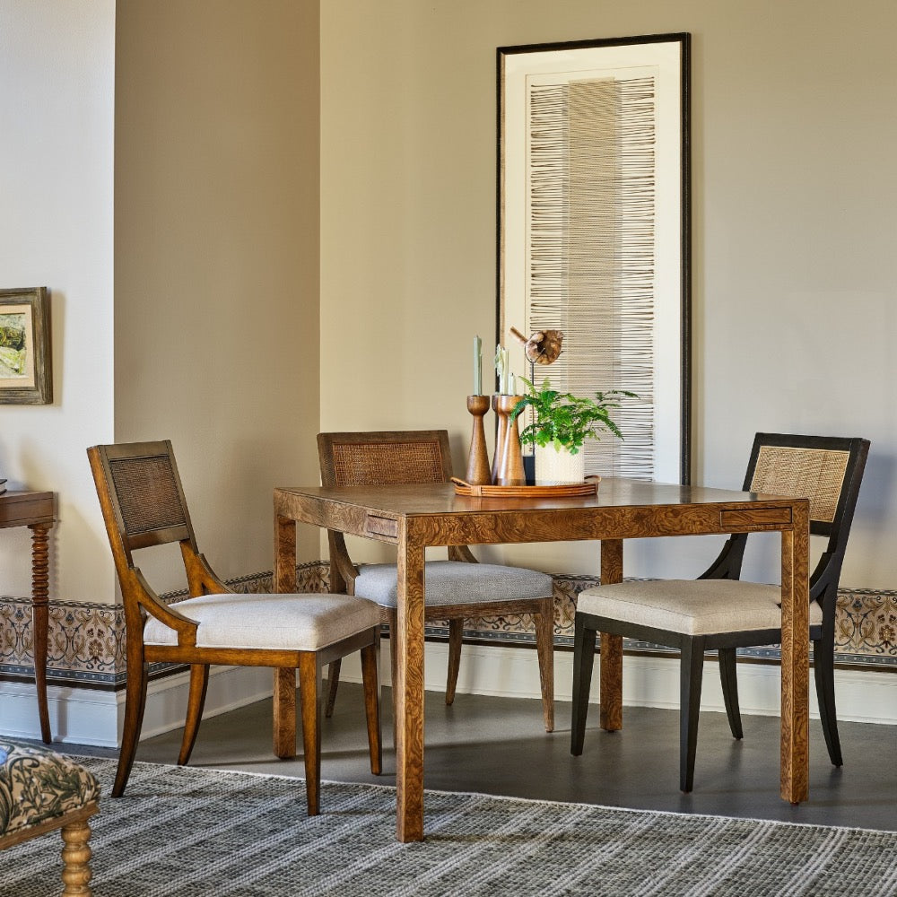 A rustic wooden dining table with four matching chairs in a styled dining room setting, featuring neutral beige walls, a framed artwork, and a patterned rug.