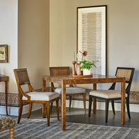 A rustic wooden dining table with four matching chairs in a styled dining room setting, featuring neutral beige walls, a framed artwork, and a patterned rug.