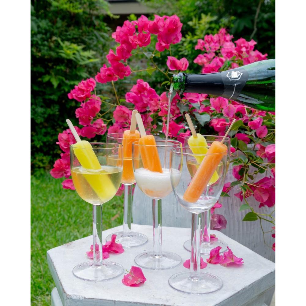 Glasses with colorful popsicles and a bottle of champagne on a table outdoors with pink flowers in the background.