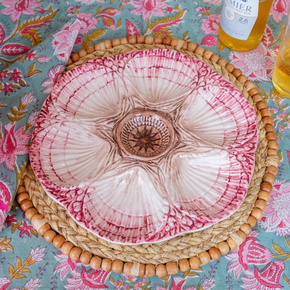 A ceramic flower-shaped serving bowl with pink and white petals and a brown center, displayed on a woven placemat over a floral tablecloth.