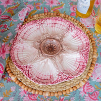 A ceramic flower-shaped serving bowl with pink and white petals and a brown center, displayed on a woven placemat over a floral tablecloth.