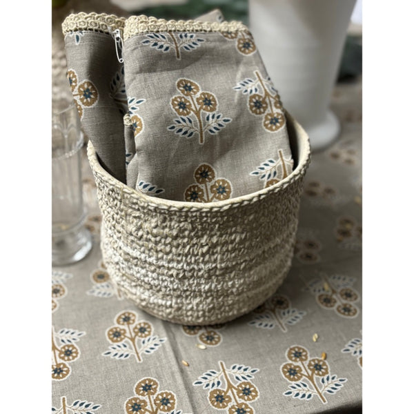 A woven basket-style pouch holding a matching fabric pouch with a floral block-print pattern in beige, brown, and blue-gray, set on a matching patterned tablecloth.