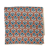 A folded square cloth tablecoth, featuring a repeating block-print style floral pattern in teal blue, off-white, and a warm terracotta orange-brown background.