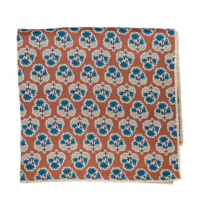 A folded square cloth tablecoth, featuring a repeating block-print style floral pattern in teal blue, off-white, and a warm terracotta orange-brown background.