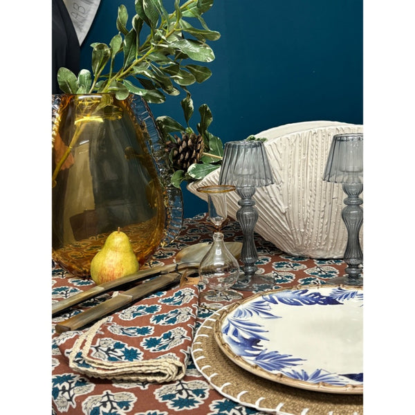 A styled dining table setting featuring a brown and blue patterned tablecloth, a blue and white floral plate, a woven napkin, a large amber glass vase with green leaves, and gray glass candlesticks and goblets against a dark teal wall.