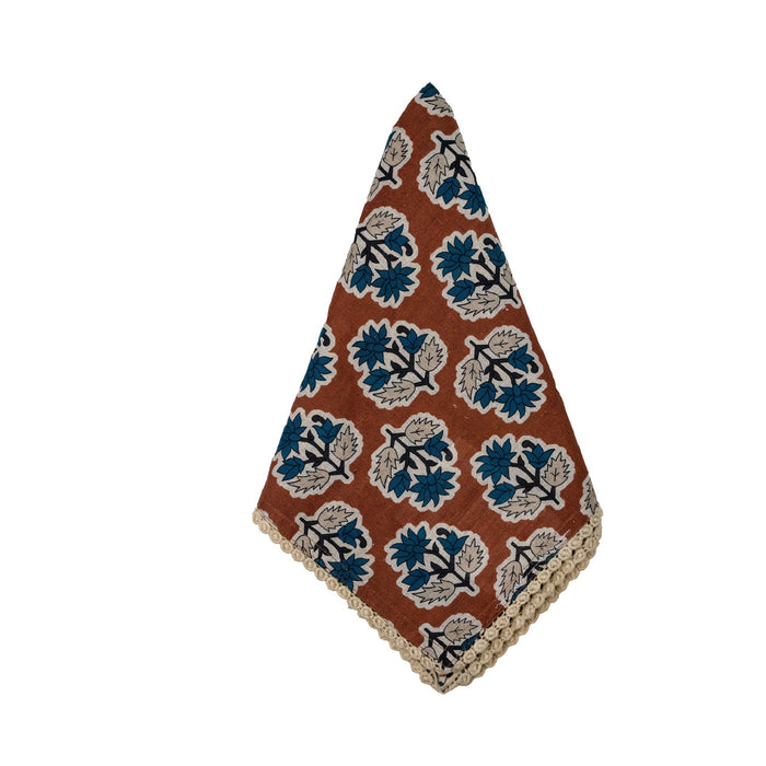 A triangular handkerchief with a rust-brown background, featuring a repeating block-print floral pattern in blue, beige, and black, finished with a decorative beige scalloped edge.