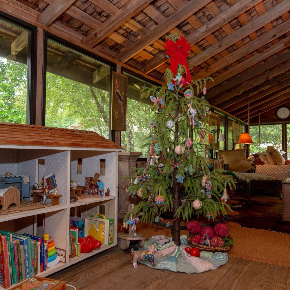 A decorated evergreen Christmas tree stands in a rustic indoor room with wood floors and large windows. The tree is adorned with colorful ornaments, a large red bow at the top, and a light fabric tree skirt.