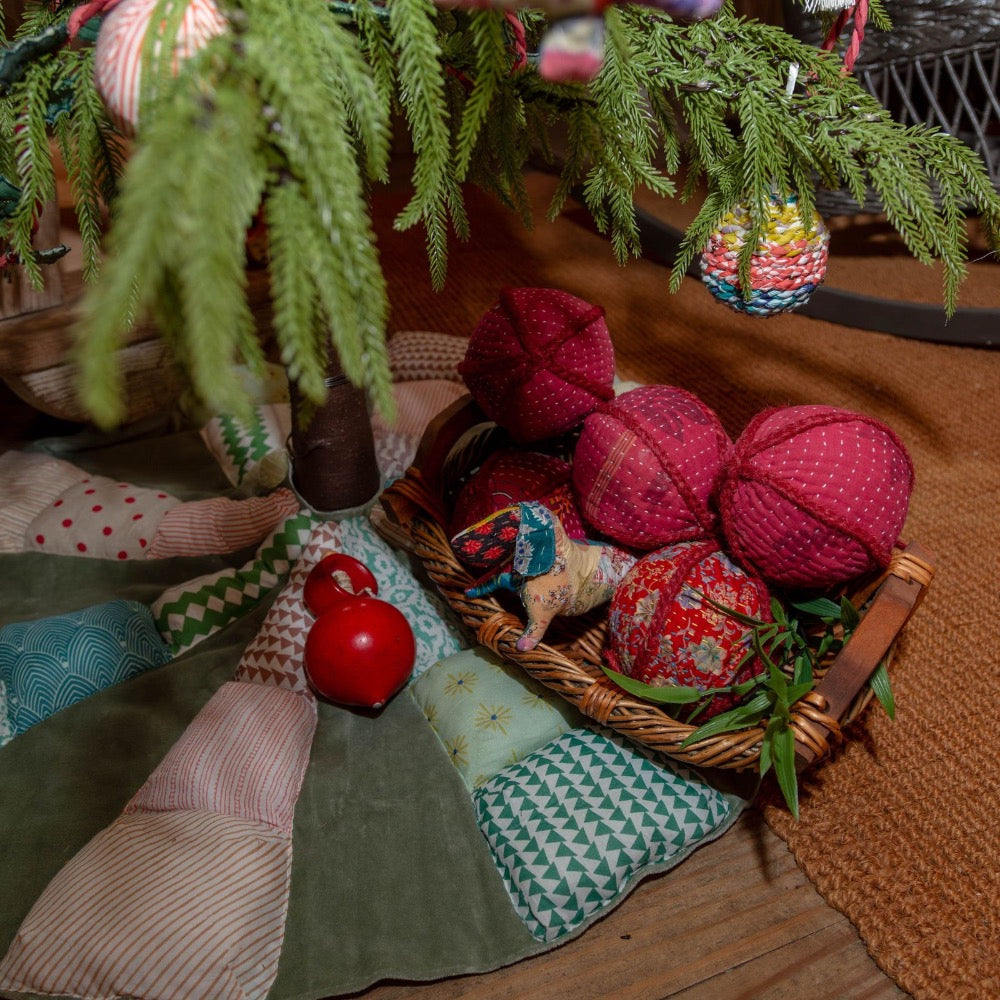 A wicker basket filled with handmade red and multicolor fabric ornaments, styled under a pine tree with a patchwork quilt and red decorative balls on a wooden floor.
