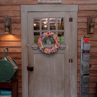 A rustic gray wooden door with a colorful crocheted wreath hanging on the front, set in a cabin-style interior with wood-paneled walls and wall-mounted lanterns.