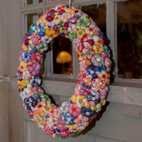 A handmade fabric flower wreath hanging on a rustic wooden door with window panes. The wreath is composed of many small, colorful fabric flowers in various patterns and colors, creating a vibrant multicolor design.
