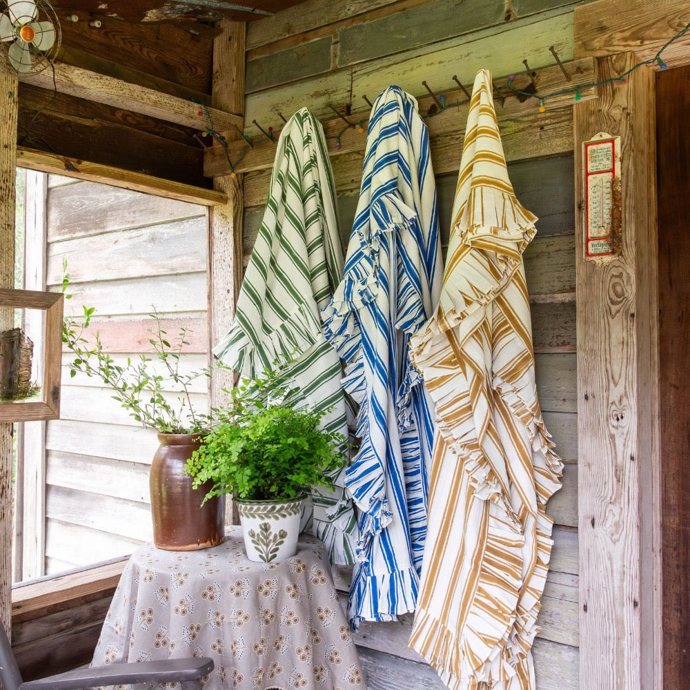 Three fringed tablecloths with vertical stripes hang on a rustic wooden wall of a porch. The blankets feature green and white stripes, blue and white stripes, and tan and cream stripes.