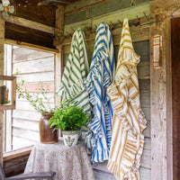Three fringed tablecloths with vertical stripes hang on a rustic wooden wall of a porch. The blankets feature green and white stripes, blue and white stripes, and tan and cream stripes.