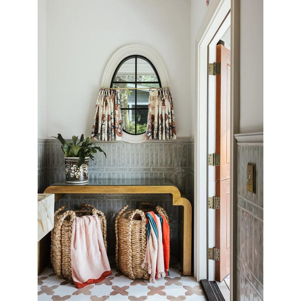A styled entryway or foyer interior with a small arched window, floral curtains, a potted plant, a wooden console table, two woven baskets filled with folded textiles, and a doorway on the right.