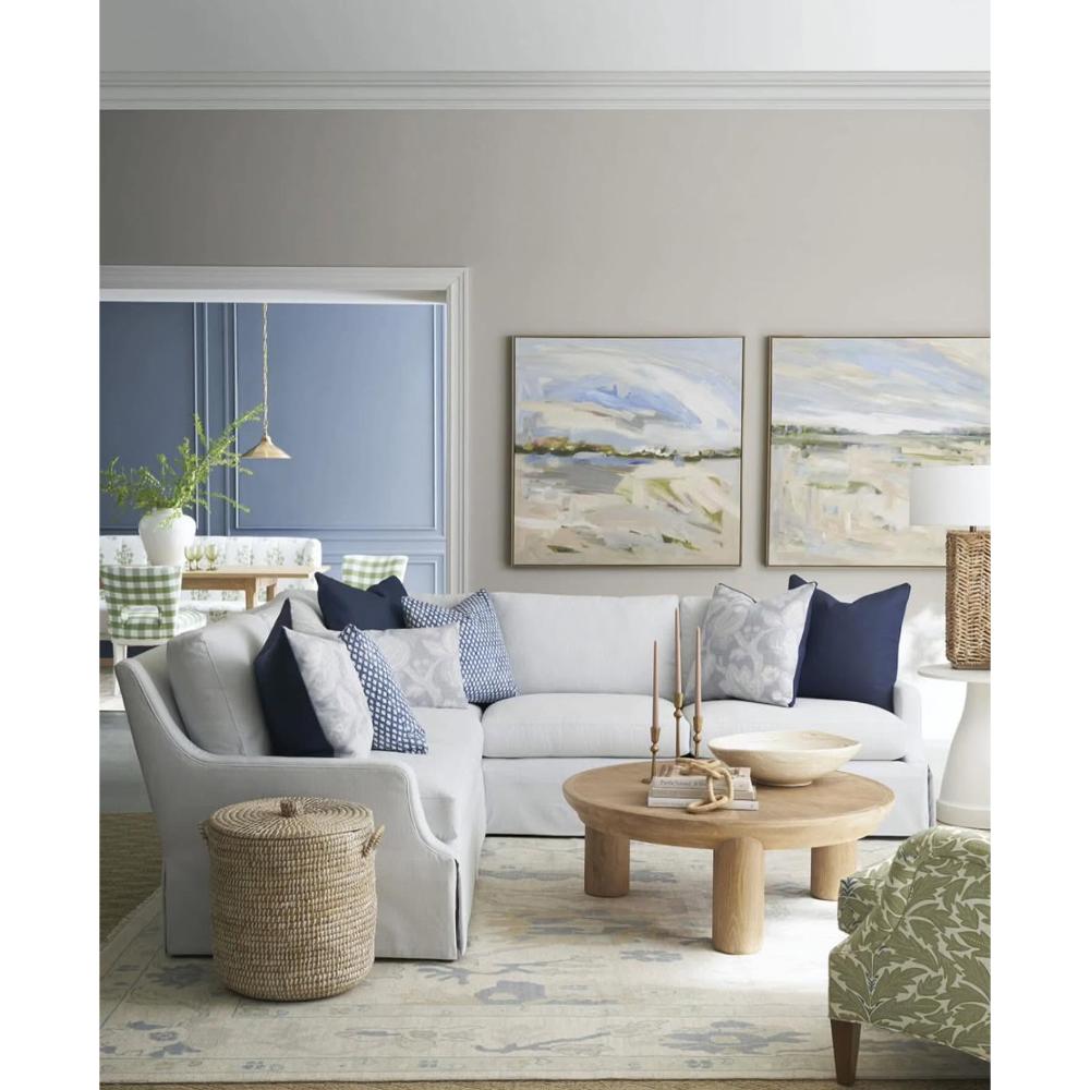 A styled living room featuring a light gray sectional sofa with navy and blue patterned pillows, a round wooden coffee table, a woven basket, and two framed landscape paintings on a gray wall.