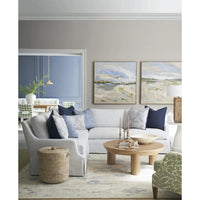A styled living room featuring a light gray sectional sofa with navy and blue patterned pillows, a round wooden coffee table, a woven basket, and two framed landscape paintings on a gray wall.