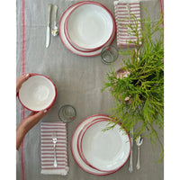 A hand places a bowl from the Always Relish Simple Round Red Rim Melamine Bowls set on a table set for two with red and white striped dishes, striped napkins, and a green plant centerpiece. Perfect for any holiday gathering.