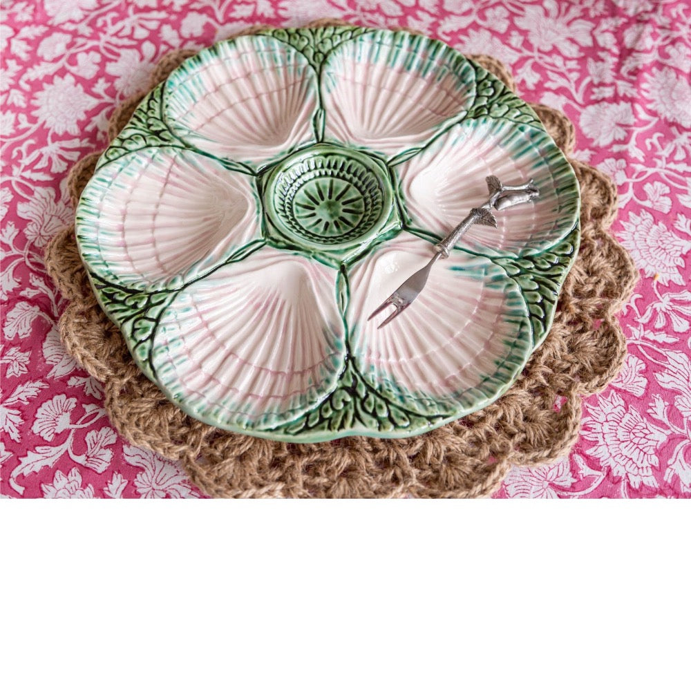 A ceramic octagonal serving platter with six shell-shaped compartments, featuring a green and white patterned glaze and a small metal fork resting in one compartment, set on a woven placemat over a pink floral fabric.
