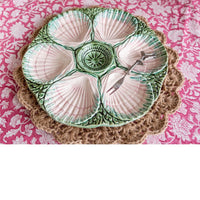 A ceramic octagonal serving platter with six shell-shaped compartments, featuring a green and white patterned glaze and a small metal fork resting in one compartment, set on a woven placemat over a pink floral fabric.
