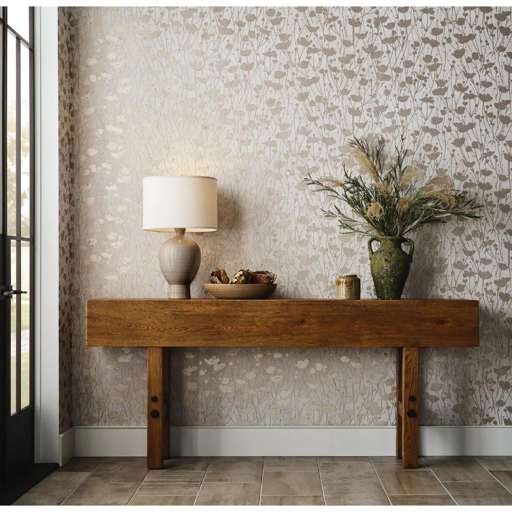 A styled interior scene showing a wooden console table against a patterned wallpaper wall, with a table lamp, bowl, candle, and vase with dried grasses on top.
