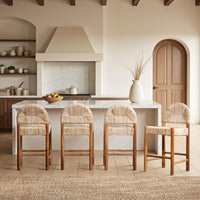 Four wooden counter stools with woven cane seats are arranged around a white kitchen island in a warm, modern kitchen setting.