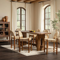 A rustic wooden dining table with six matching counter stools featuring woven straw seats, set on a patterned area rug in a bright, modern farmhouse-style dining room.