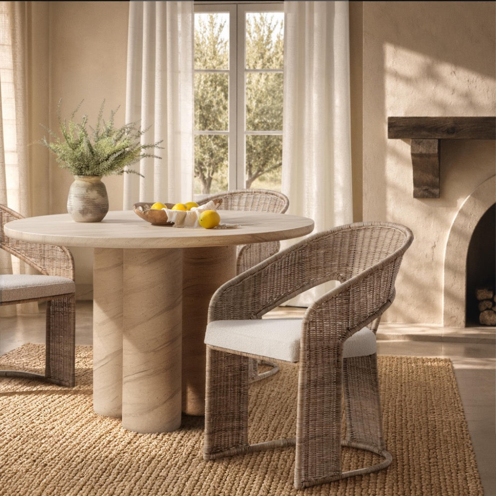 A round dining table with a light stone top and cylindrical pedestal base, paired with woven wicker chairs featuring white cushions, set in a warm, sunlit room with a window, curtains, rug, and fireplace.