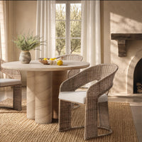 A round dining table with a light stone top and cylindrical pedestal base, paired with woven wicker chairs featuring white cushions, set in a warm, sunlit room with a window, curtains, rug, and fireplace.