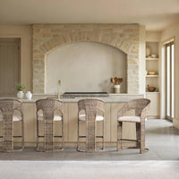Four woven wicker bar stools with cream cushions arranged around a light wood kitchen island in a warm, modern stone kitchen interior.