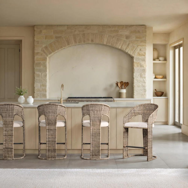 Four woven wicker bar stools with cream cushions arranged around a light wood kitchen island in a warm, modern stone kitchen interior.