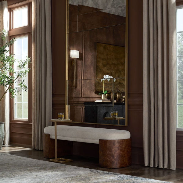 A large rectangular wall mirror with a gold frame hangs in a richly decorated room. The mirror reflects a dark console table and decor, while the room itself features warm brown walls, beige curtains, a matching upholstered bench with textured brown legs, and tall windows with natural light.