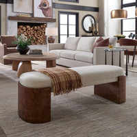 A modern upholstered bench with a cream fabric seat and dark brown cylindrical block legs, styled in a living room with a plaid throw blanket draped over it.