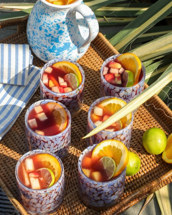Cocktail glasses with fruit and a pitcher on a woven tray outdoors.
