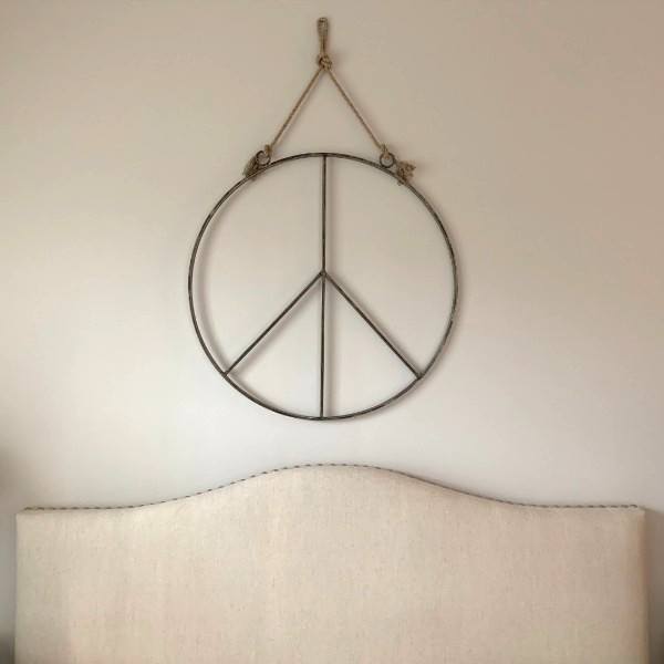 Sugarboo & Co. Hanging Metal + Jute Peace Sign hangs above a light headboard against a plain wall.