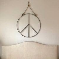 Sugarboo & Co. Hanging Metal + Jute Peace Sign hangs above a light headboard against a plain wall.