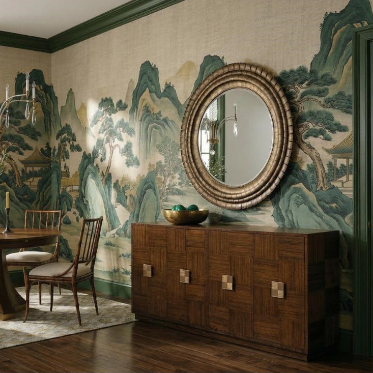 A styled dining room interior featuring a round wooden dining table with four matching wooden chairs, a large round decorative mirror on a wooden sideboard, and a wall mural with green and blue mountain scenery.