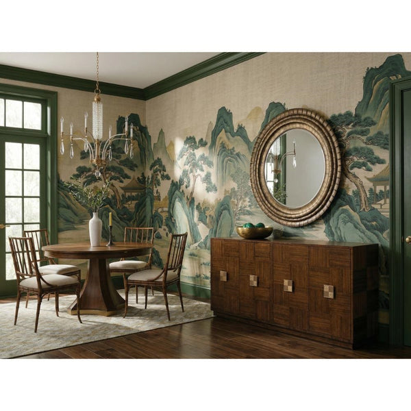 A styled dining room interior featuring a round wooden dining table with four matching wooden chairs, a large round decorative mirror on a wooden sideboard, and a wall mural with green and blue mountain scenery.
