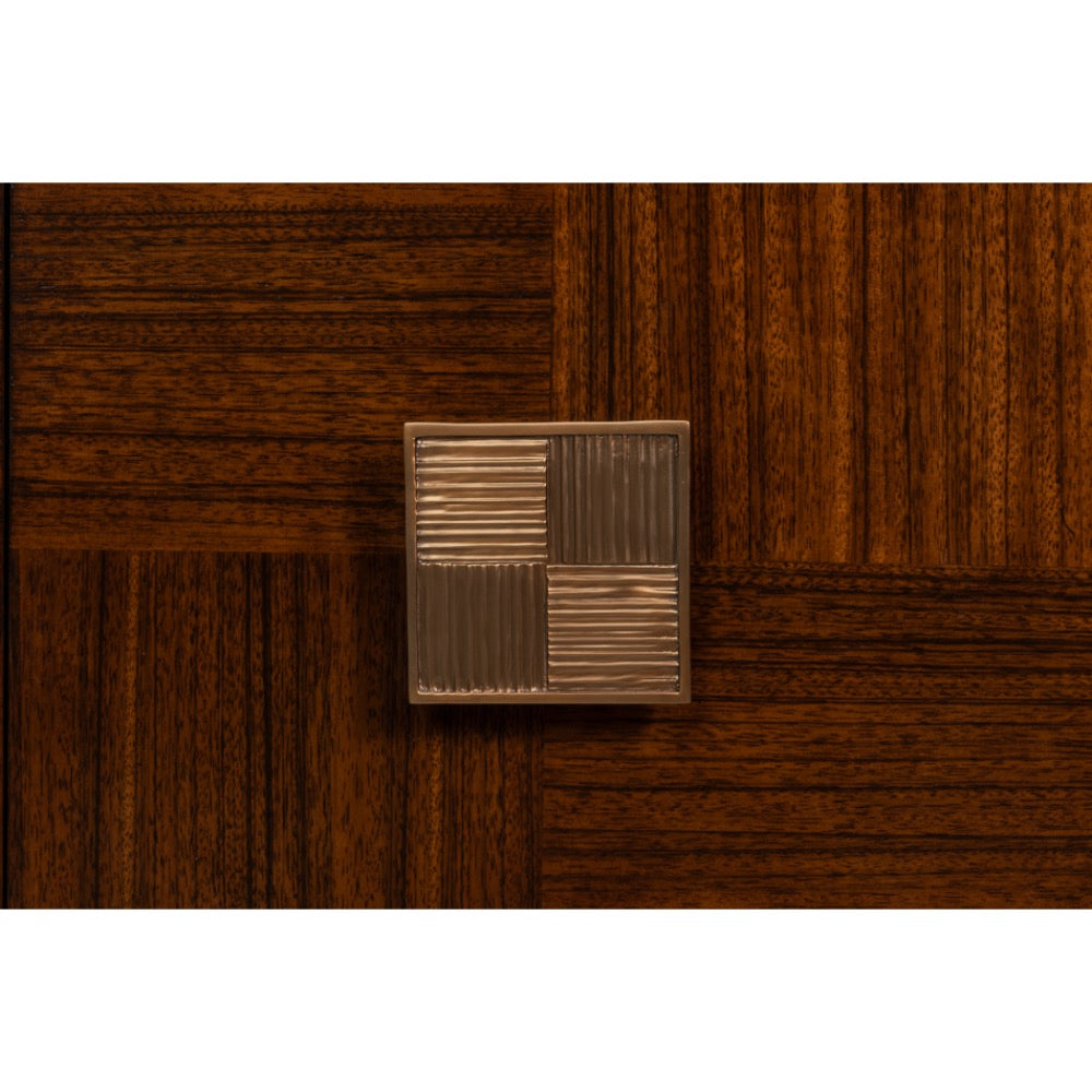 A square metallic cabinet knob with a textured four-quadrant design, mounted on a dark brown wood parquet background.