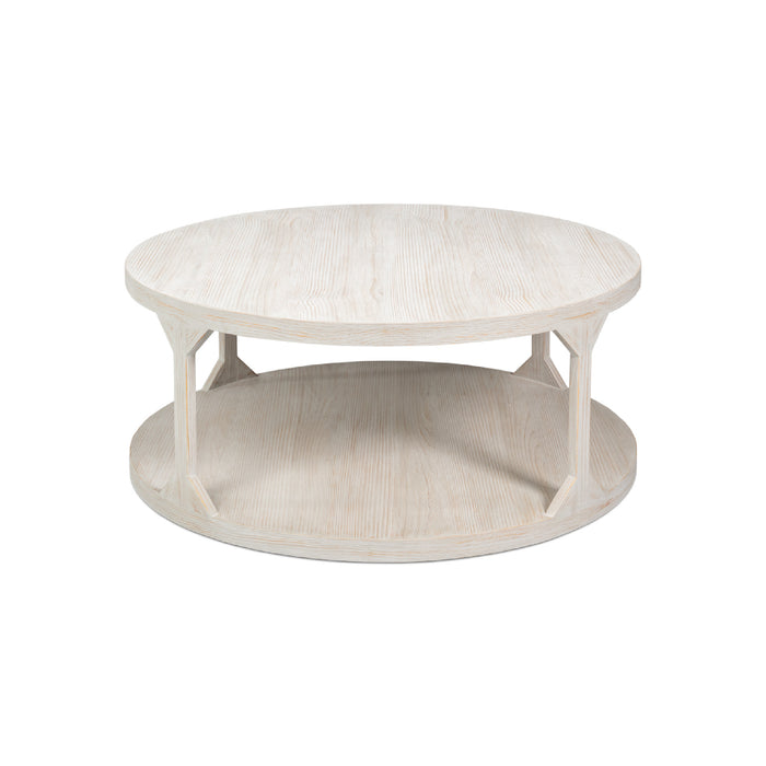 A round, low-profile coffee table with a whitewashed light wood finish, featuring a top surface, a lower shelf, and four angled support legs.