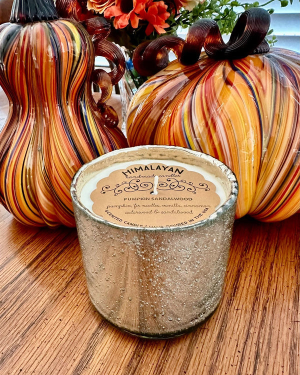 A candle in a textured metallic silver cup sits on a wooden table, styled with decorative glass pumpkins in the background. The candle label reads 'HIMALAYAN Candle Company' and 'Pumpkin Sandalwood'.