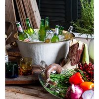 Green bottles in a beverage tub filled with ice, surrounded by vegetables and a plant on a rustic wooden table.