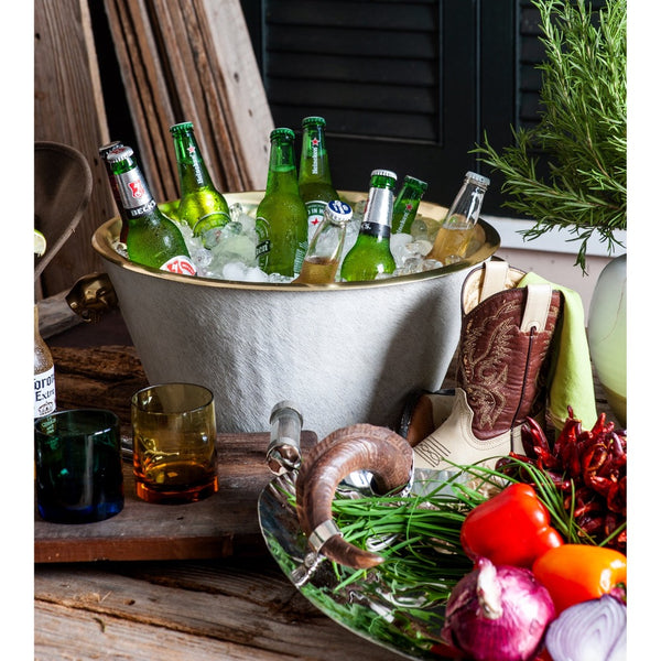 Green bottles in a beverage tub filled with ice, surrounded by vegetables and a plant on a rustic wooden table.