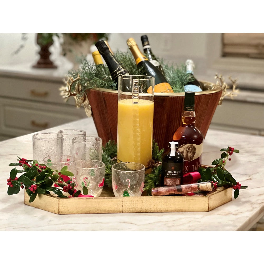 A holiday-themed beverage arrangement on a marble countertop, featuring a wooden bucket filled with greenery and bottles, a tall glass of orange juice, several textured drinking glasses, a bottle of liquor, and holiday greenery with red berries on a wooden tray.