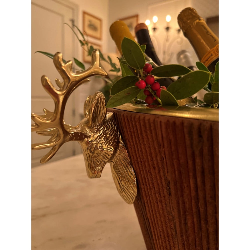 A close-up of a decorative gold reindeer head ice bucket with a ribbed brown wooden body, styled with green leaves and red berries, with champagne bottles partially visible in the background.