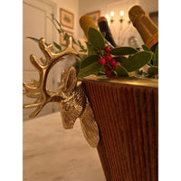 A close-up of a decorative gold reindeer head ice bucket with a ribbed brown wooden body, styled with green leaves and red berries, with champagne bottles partially visible in the background.