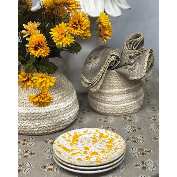 A styled tabletop scene with woven storage baskets, folded patterned fabric napkins, a vase of yellow flowers, and a stack of white plates with yellow speckles on a patterned tablecloth.